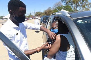 A masked man gives a shot to a masked person sitting in a car.