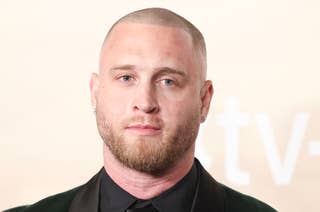 Chet Hanks with a shaved head and beard wearing a velvet suit jacket and black shirt stands in front of a backdrop with the Apple TV+ logo