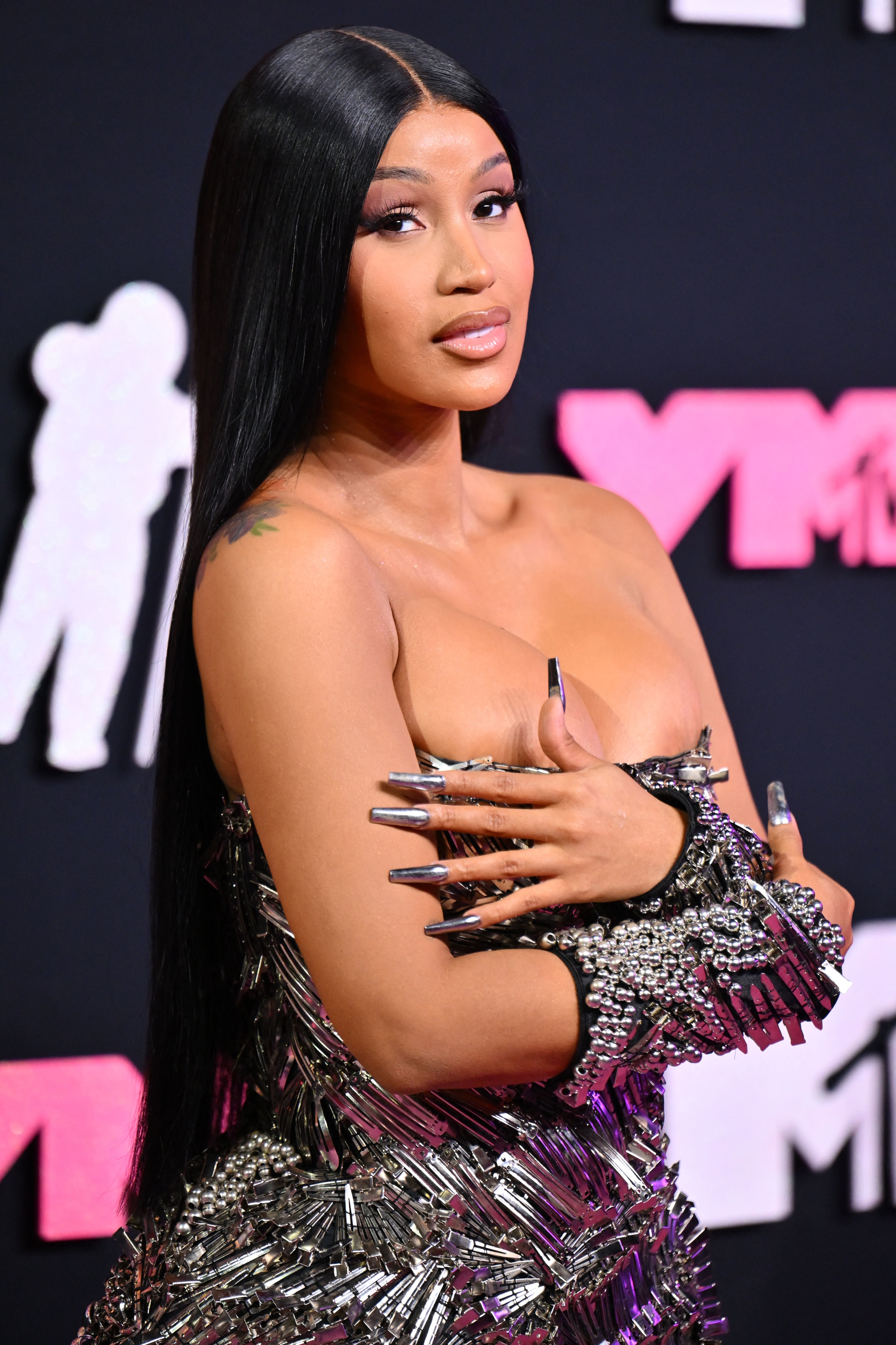 Cardi B on the red carpet wearing a strapless, metallic gown adorned with silver spiked detailing. She has long, straight hair and is holding her arms across her chest