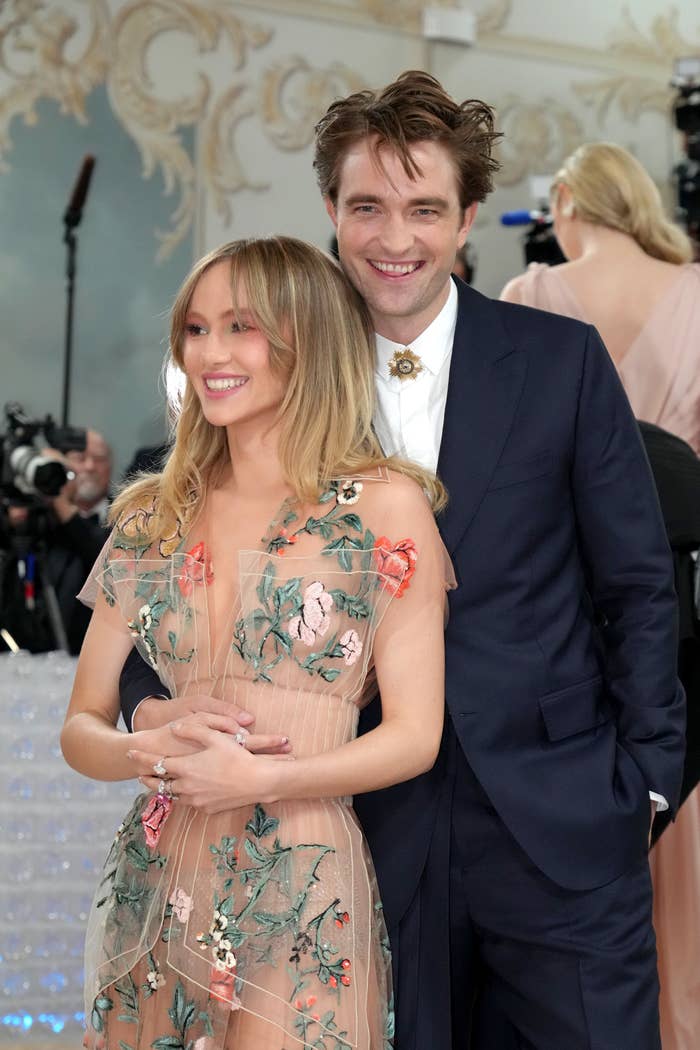 Suki Waterhouse and Robert Pattinson smile and pose together at a formal event. Suki wears a floral embroidered dress, and Robert wears a dark suit with a shirt