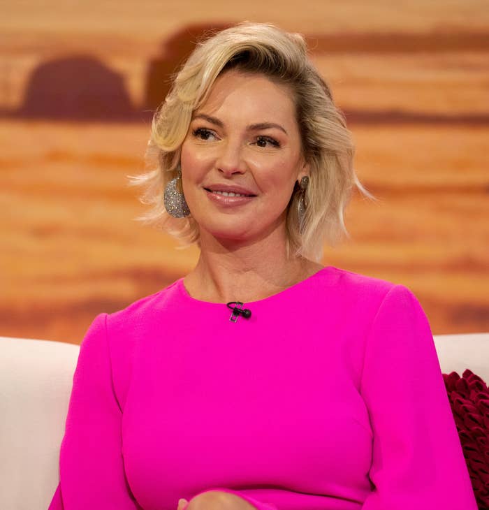 Katherine Heigl sits on a couch, wearing a bright dress, smiling during an interview