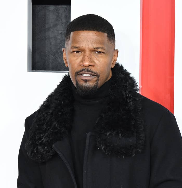 Jamie Foxx on a red carpet, wearing a stylish long dark coat with a fur collar over a dark-colored turtleneck