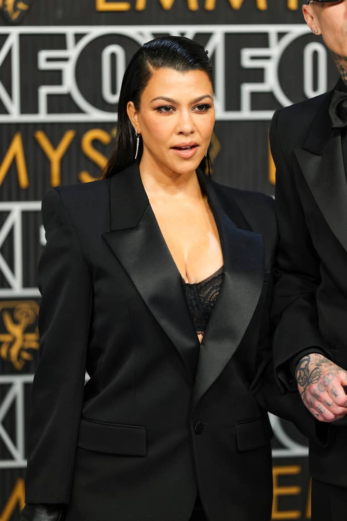 Kourtney Kardashian on a red carpet, wearing a sleek black blazer over a low-cut top. She is standing next to a person with tattoos on their hand