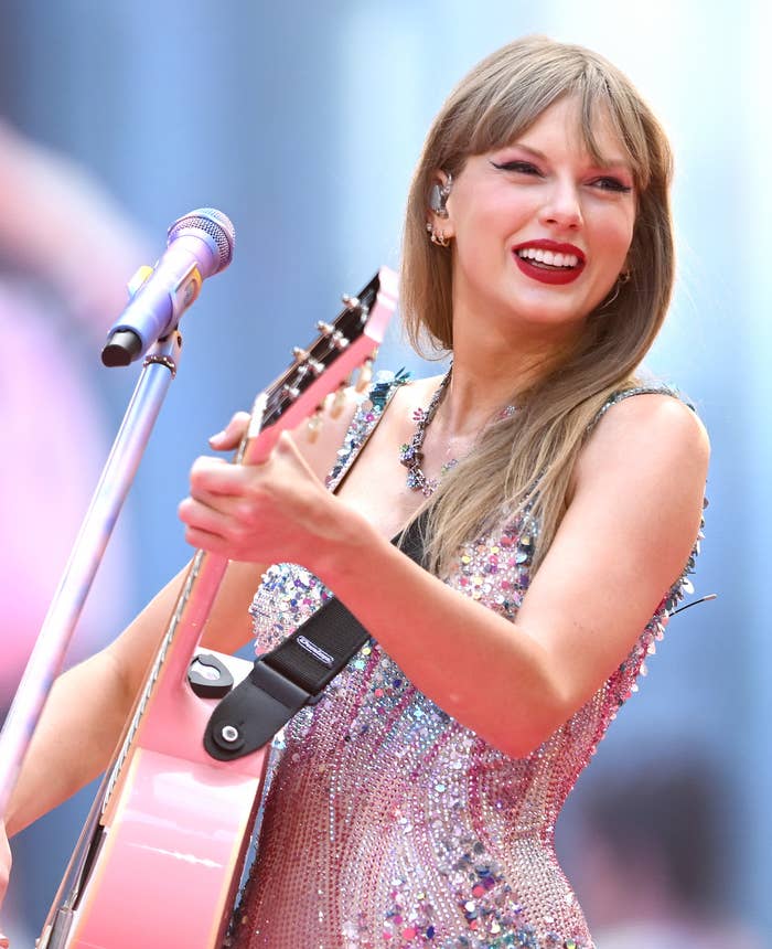 Taylor Swift performs on stage with a pink guitar, wearing a sparkly, colorful bodysuit. She is smiling and appears animated during her performance