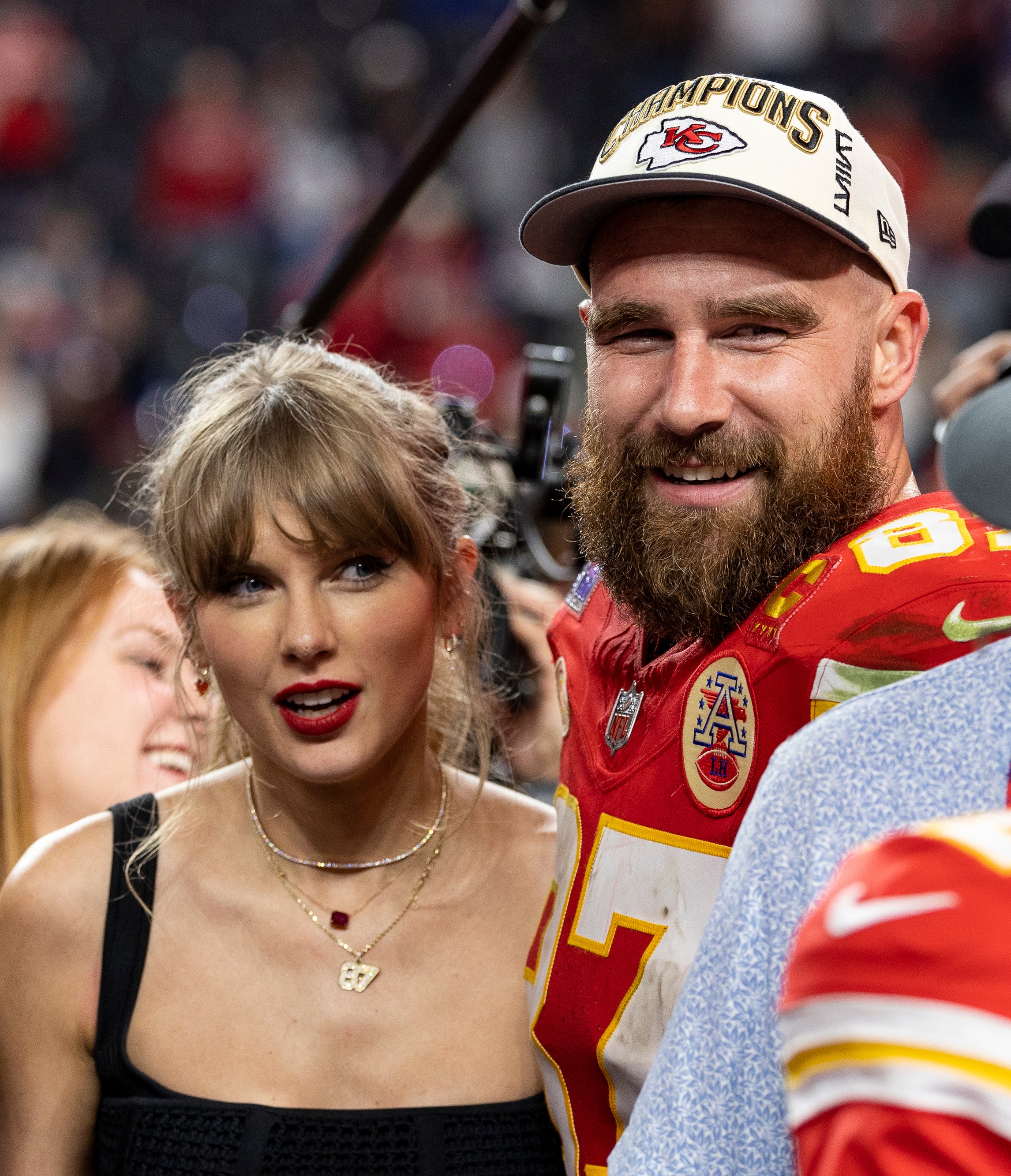 Taylor Swift and Travis Kelce smiling together surrounded by people and media cameras. Travis is in a football uniform and championship cap, standing near Mecole Hardman