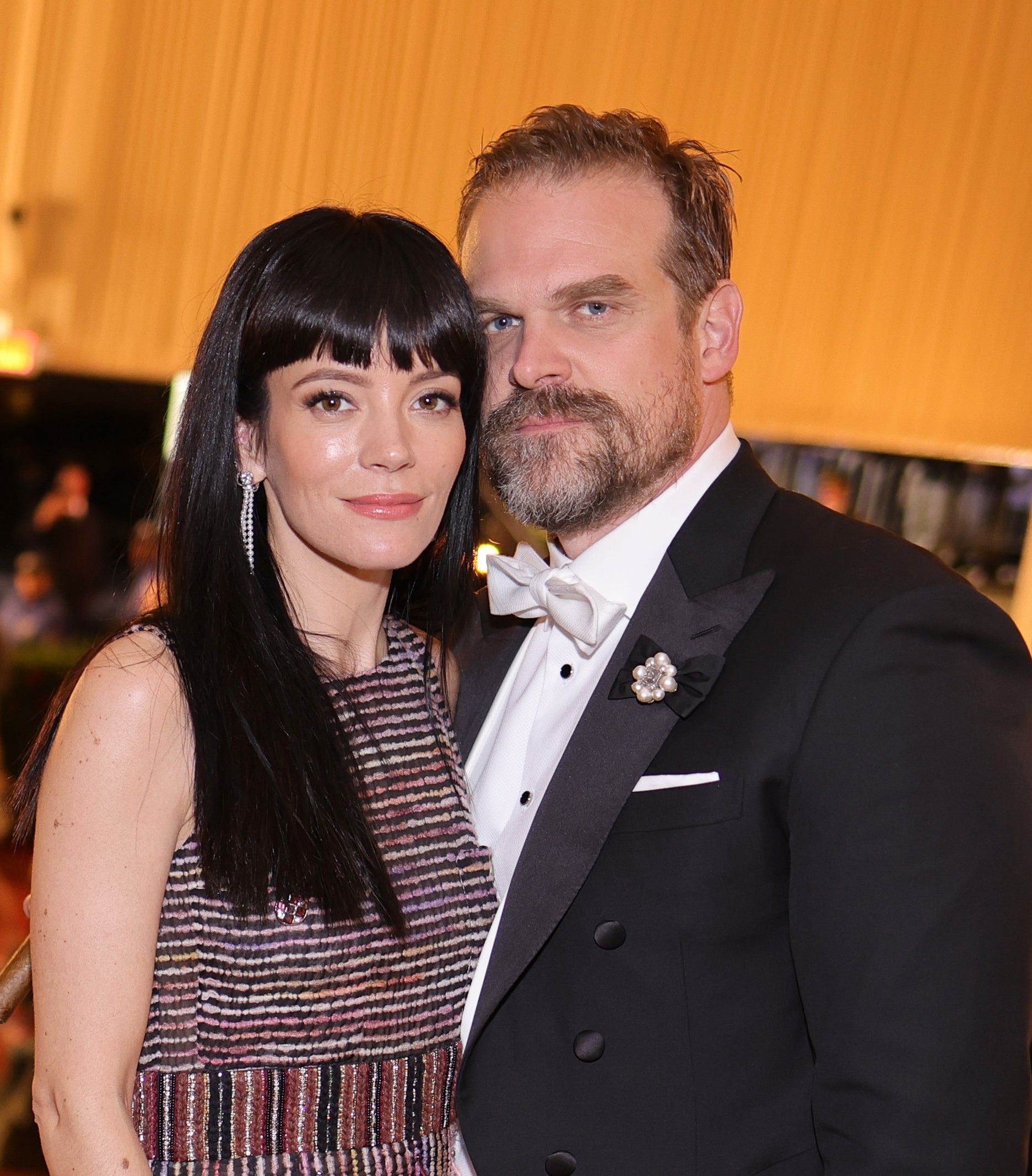 Lily Allen, wearing a fringed dress, and David Harbour, in a tuxedo, pose together at a formal event. He holds a hat in his left hand