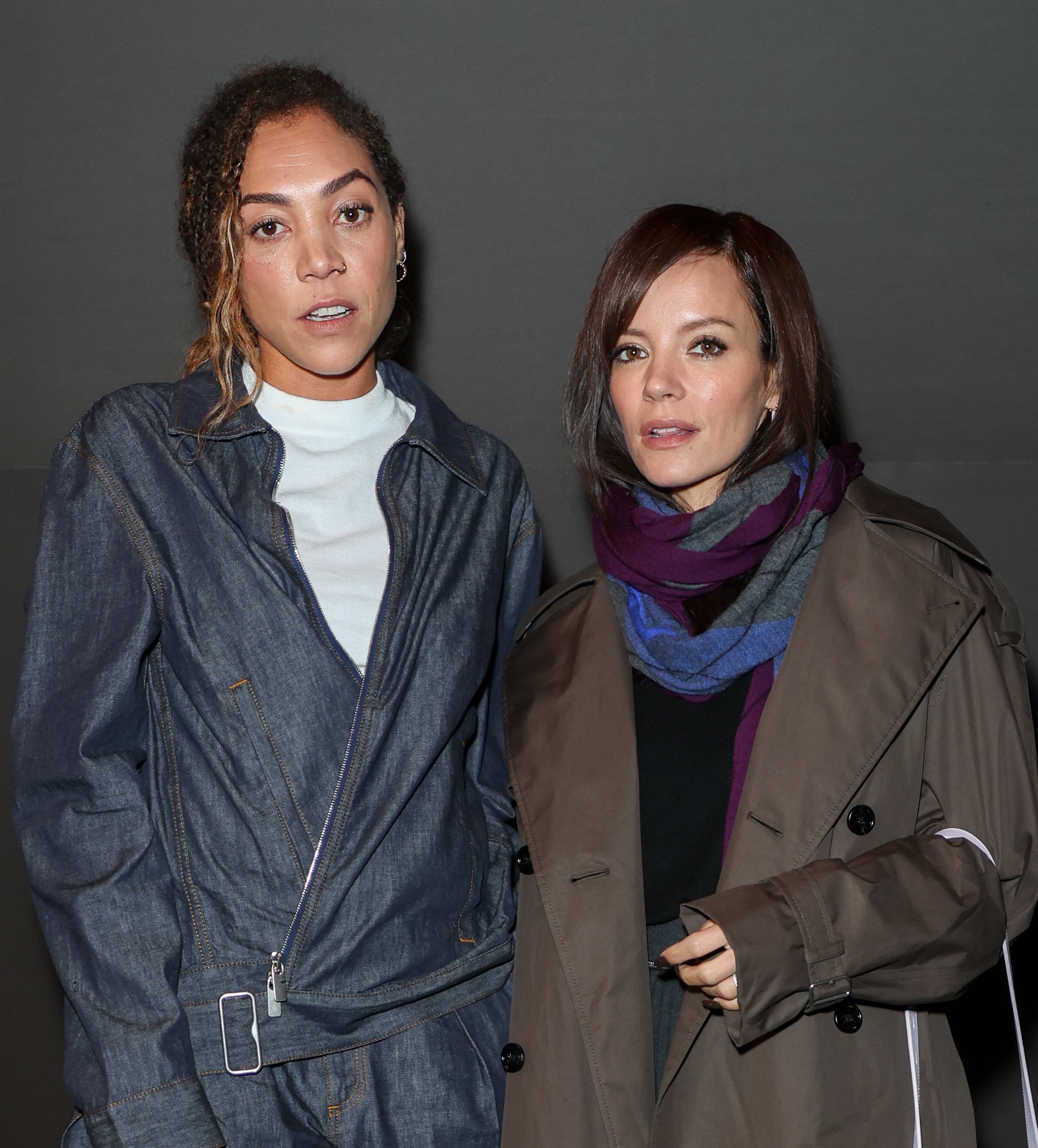 Miquita Oliver in a denim jacket and pants with a shoulder bag, stands next to Lily Allen in a long coat with a fur bag, both posing for a photo