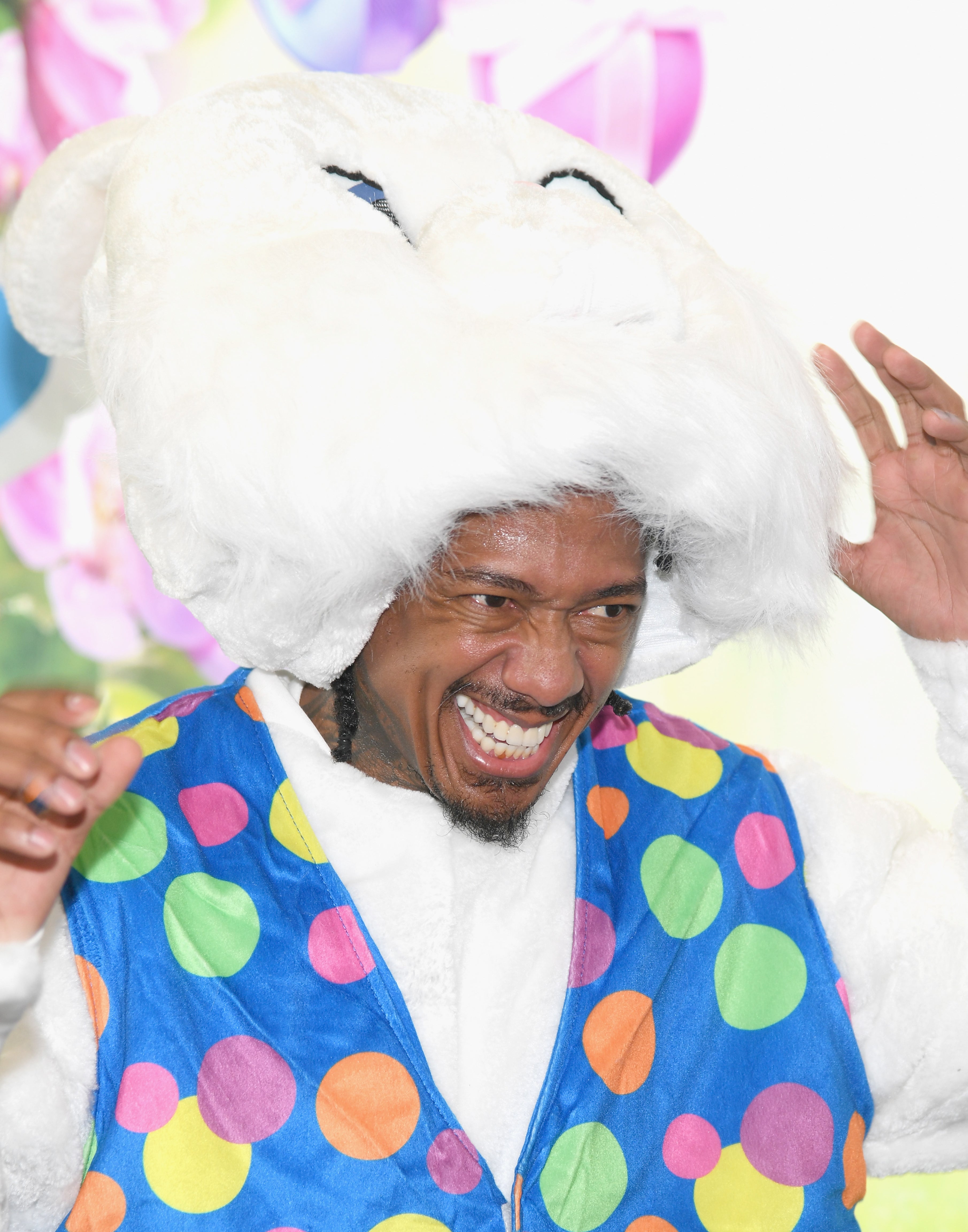 Nick Cannon dressed in a colorful polka dot vest and a large fluffy bunny hat, smiling and holding the hat