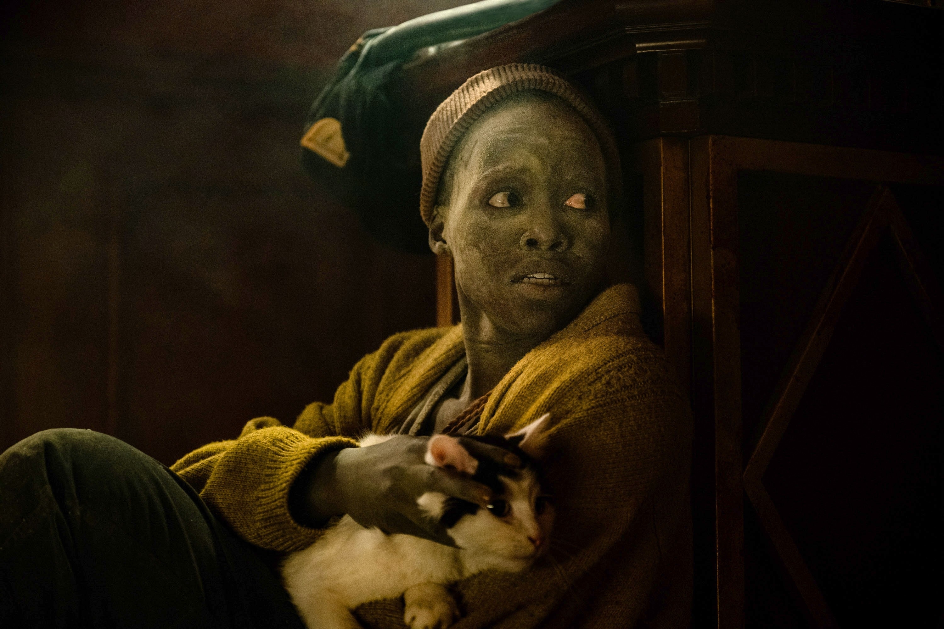 Lupita Nyong&#x27;o with distressed expression, covered in dark makeup, wearing a knitted beanie and cardigan, holding a white and black cat