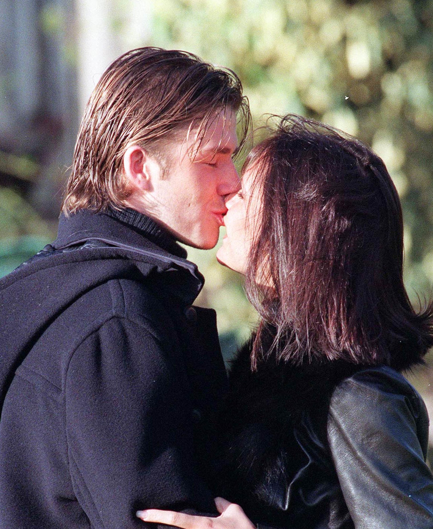 Victoria and David Beckham embrace and kiss outdoors in 1998. Both are wearing dark coats