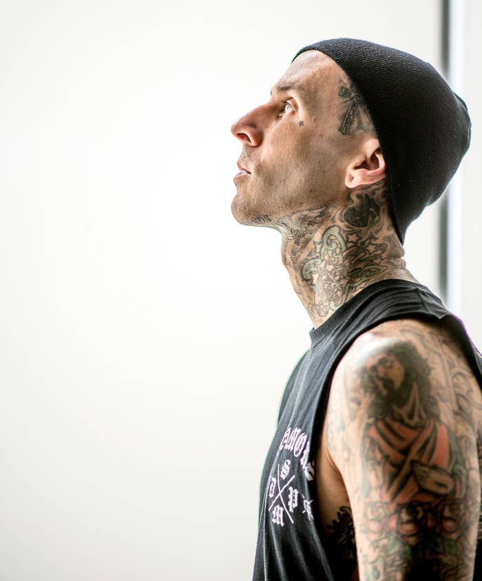 Travis Barker looks upward, wearing a black beanie and a sleeveless black shirt, showcasing his heavily tattooed neck and arms