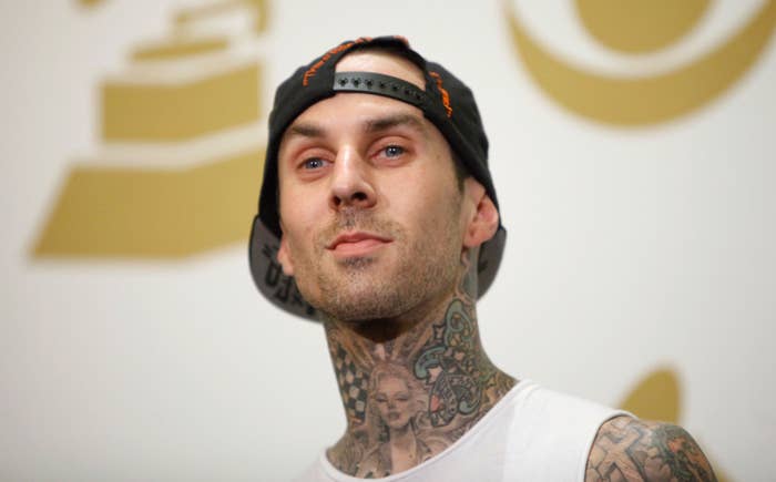 Travis Barker, wearing a backward cap and white tank top, stands with visible tattoos on his neck, in front of a backdrop with music award symbols