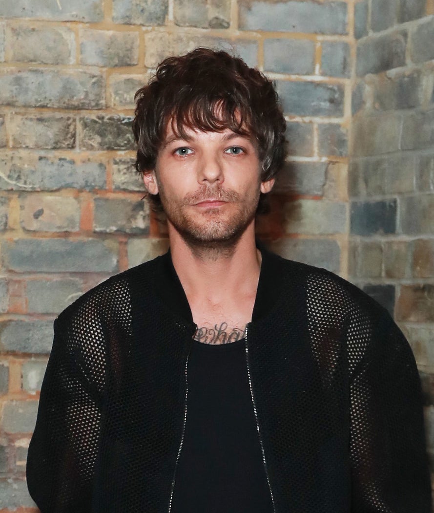 Louis Tomlinson stands against a brick wall, wearing a black mesh jacket, black shirt and black pants