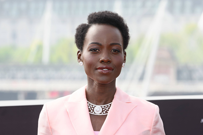 Lupita Nyong'o poses outdoors in a light-colored suit with a croc-styled necklace and earrings