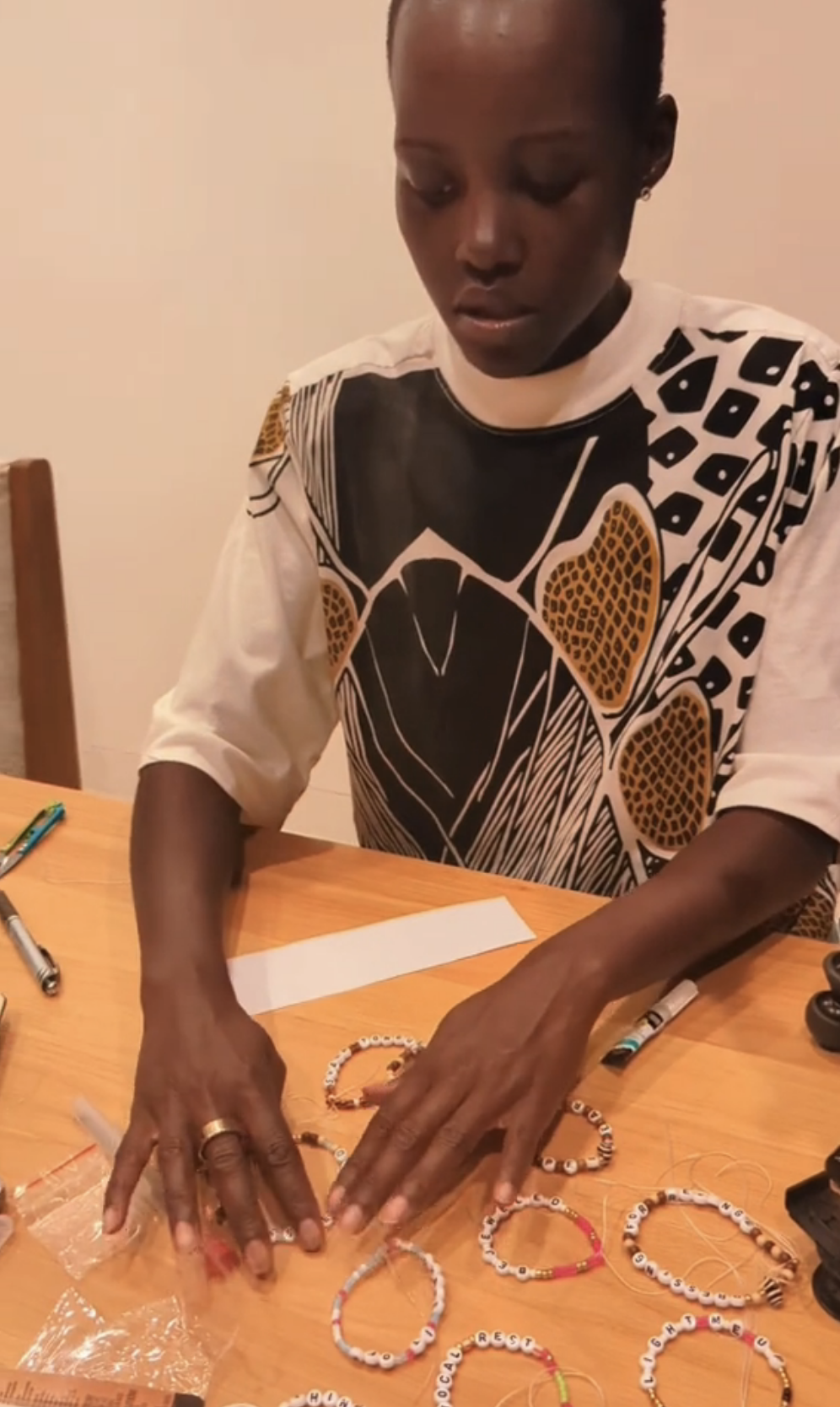 Lupita Nyong&#x27;o sits at a table arranging beaded bracelets. She wears a patterned shirt and is focused on her work. Markers and materials are on the table